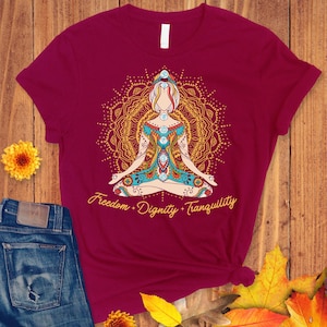 May include: A maroon t-shirt featuring a seated figure in a yoga pose, with a colorful design and a golden mandala background. The text "Freedom + Dignity + Tranquility" is written in a script font. The shirt is paired with blue jeans.