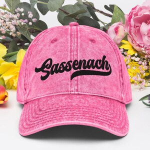 May include: A pink baseball cap featuring the word "Gassenach" in black script. The cap has a faded, vintage appearance and a curved peak. The background includes flowers and foliage.