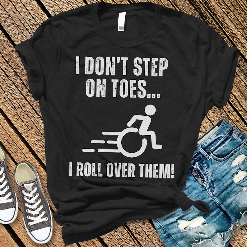 May include: Black t-shirt with white text that reads "I DON'T STEP ON TOES... I ROLL OVER THEM!" A graphic of a person in a wheelchair is also printed on the shirt. The shirt is paired with denim shorts and gray sneakers.