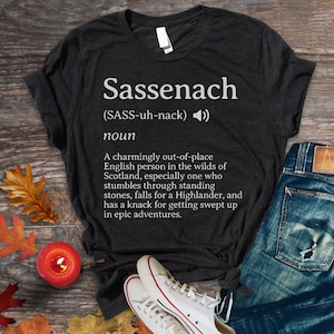 Könnte beinhalten: Dunkelgraues T-Shirt mit dem Wort "Sassenach" in Weiß und seiner Definition. Das Shirt wird mit blauen Jeans und weißen Turnschuhen kombiniert. Herbstblätter und eine Kerze sind im Hintergrund.