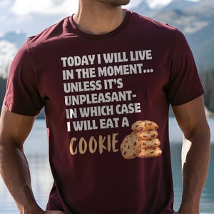 Könnte beinhalten: Burgunderfarbenes T-Shirt mit dem Text "TODAY I WILL LIVE IN THE MOMENT... UNLESS IT'S UNPLEASANT- IN WHICH CASE I WILL EAT A COOKIE" in Weiß und Gold. Ein Stapel Schokoladenkekse ist darunter abgebildet.