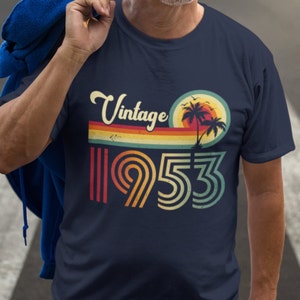 Puede incluir: Una camiseta azul marino con un diseño vintage de 1953. El diseño presenta una puesta de sol con palmeras y una franja de arcoíris.