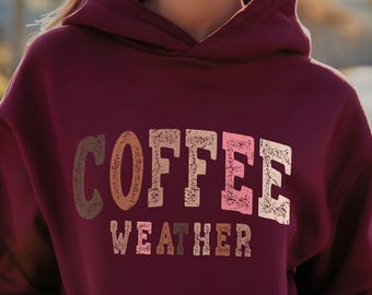 Puede incluir: Una sudadera con capucha color burdeos con el texto "COFFEE WEATHER" en una fuente de estilo vintage con colores marrón, rosa y naranja.