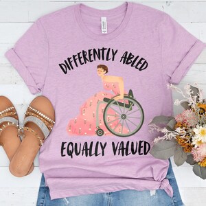 Peut inclure: Un t-shirt lilas avec l'inscription "DIFFERENTLY ABLED EQUALLY VALUED" en noir. Le t-shirt présente une illustration d'une personne en fauteuil roulant portant une robe rose à motifs floraux. Des sandales et des fleurs sont également visibles.