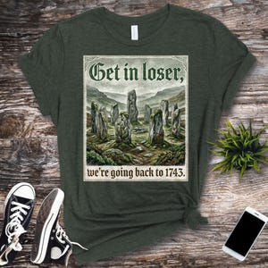 Puede incluir: Camiseta verde oscuro con el texto "Get in loser, we're going back to 1743." El gráfico muestra un paisaje con piedras verticales. La camiseta se combina con zapatillas deportivas blancas y negras.