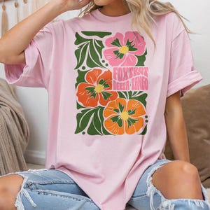 Könnte beinhalten: Hellrosa T-Shirt mit floralem Design und dem Text "FOXTROT DELTA TANGO". Das Design zeigt rosa und orangefarbene Blumen mit grünen Blättern. Das T-Shirt ist aus weichem Material.