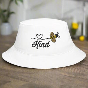 Op de afbeelding: Witte vissershoed met zwart geborduurd ontwerp. Het ontwerp bevat het woord "Kind", een hart en een bij. De hoed is gemaakt van zacht materiaal, geschikt voor casual wear.