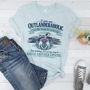 Puede incluir: Una camiseta jaspeada de color azul claro con un estampado gráfico de un castillo escocés y un cardo morado. El texto dice: I am an OUTLANDERAHOLIC on the road to recovery. Just kidding, I'm on my way to WATCH ANOTHER EPISODE.