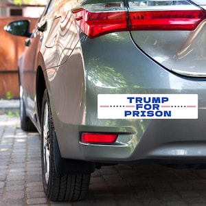 May include: A gray car with a bumper sticker that reads "TRUMP FOR PRISON" with red, white, and blue stars.