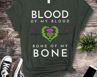 May include: A dark green t-shirt with white text that reads "Blood of my blood, bone of my bone". A stylized green thistle with a purple blossom is centered on the shirt.