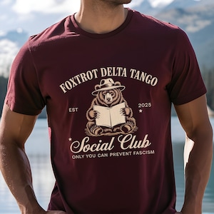 Könnte beinhalten: Burgunderfarbenes T-Shirt mit einem Bären, der einen Hut trägt und ein Buch liest. Auf dem Shirt steht "Foxtrot Delta Tango Social Club" und "Only You Can Prevent Fascism". Das Jahr "2025" ist ebenfalls aufgedruckt.