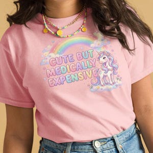 May include: Pink t-shirt with the text "Cute But Medically Expensive" above a rainbow and a unicorn illustration. The unicorn has a flowing mane and tail in shades of pink, purple, and blue.