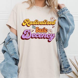 Puede incluir: Una camiseta beige con un gráfico de estilo retro que dice "Radicalizado por la decencia básica". El gráfico está en tonos de amarillo, naranja, morado y rojo con flores blancas y brillos.