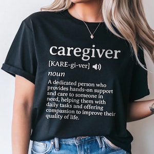May include: Black t-shirt with the word "caregiver" in white, along with a definition. The definition describes a dedicated person providing hands-on support and care. The shirt also includes the phonetic pronunciation of the word.