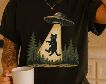 May include: Black t-shirt featuring a graphic of a black cat being abducted by a UFO. The design includes a forest backdrop and a beam of light shining down on the cat. The UFO is gray and the cat has a surprised expression.
