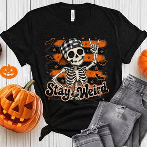 Può includere: Una t-shirt nera con la grafica di uno scheletro che indossa un berretto da baseball a quadri bianchi e neri, facendo un gesto rock-and-roll con la mano. Il design include pennellate arancioni, piccole stelle, pipistrelli e il testo Stay Weird in nero.
