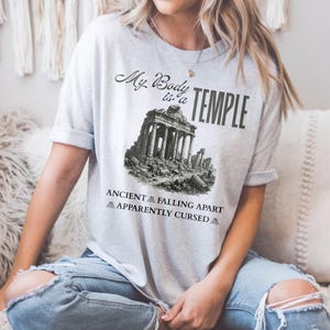 May include: A light gray t-shirt with the text "My Body is a Temple" above a black and white illustration of an ancient temple. Below the illustration are the words "Ancient Falling Apart Apparently Cursed".