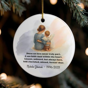 Può includere: Ornamento natalizio rotondo in ceramica bianca, con un cordino dorato per appenderlo. Presenta un'illustrazione di una madre e un bambino con le ali d'angelo, e il testo "Those we love never truly part..." e il nome "Nicholas Zahorcak - 1996-2021."
