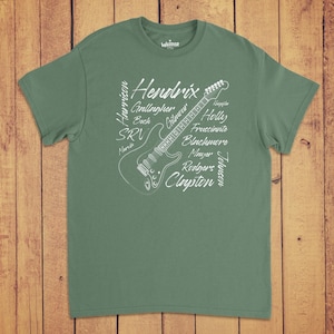 Pode incluir: Uma t-shirt de manga curta verde s&aacute;lvia com um gr&aacute;fico de linhas brancas de uma guitarra el&eacute;trica. O design inclui os nomes Hendrix, Harrison, Gallagher, Beck, SRV, Marvin, Gilmour, Holly, Frusciante, Blackmore, Mayer, Rodgers, Clayton, Knopfler e Johnson.
