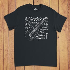 Pode incluir: Uma t-shirt preta de manga curta com um gr&aacute;fico branco de uma guitarra el&eacute;trica rodeada pelos nomes de guitarristas famosos como Hendrix, Gallagher, Beck, SRV, Gilmour, Holly, Frusciante, Blackmore, Mayer, Rodgers, Johnson e Clapton.
