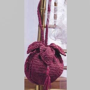 May include: A burgundy crochet drawstring bag with a matching strap and tassel. The bag has a rounded shape with a textured surface. The strap is attached to the top of the bag, and the tassel hangs from the bottom.