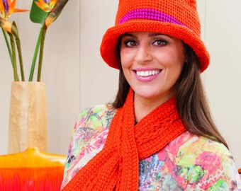May include: A woman wearing a bright orange crocheted hat with a purple band and a matching orange crocheted scarf. The hat has a wide brim and the scarf is long and flowing.