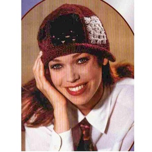 Op de afbeelding: Een vrouw draagt een rode, zwarte en witte gehaakte hoed met een brede rand. De hoed heeft een geometrisch patroon en wordt licht schuin gedragen. Ze glimlacht en heeft lang, donker haar.