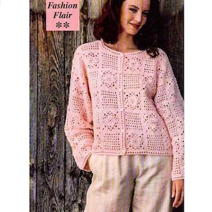 May include: A pink crocheted cardigan with a button-up front and a square pattern. The cardigan is worn by a person with brown hair and is photographed against a wood background.