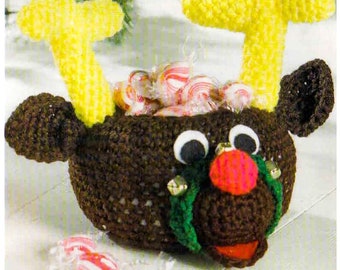 May include: A brown crocheted reindeer basket with yellow antlers, green holly, and red bells. The basket is filled with candy canes.