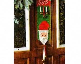 Può includere: Un appendino per porta di Natale verde e rosso con un viso di Babbo Natale e le parole "HO HO HO" in bianco. L'appendino ha un fiocco bianco con campane in fondo.