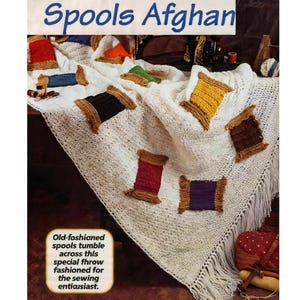 May include: A white crocheted afghan with colorful yarn spool appliqués. The spools are in various colors, including red, yellow, green, and purple. The afghan has a fringed edge and text that reads "Spools Afghan".