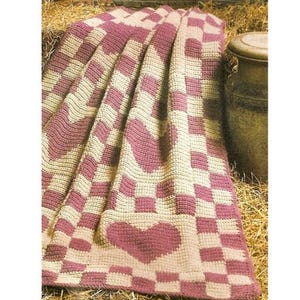 Peut inclure: Une couverture faite à la main, crochetée, avec un motif en damier crème et mauve. La couverture présente de grands cœurs au centre et sur les bords. La couverture est pliée et drapée sur une surface.