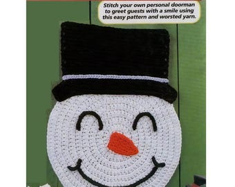 Può includere: Applique da parete a forma di pupazzo di neve all'uncinetto con cappello a cilindro nero e volto sorridente. Il volto del pupazzo di neve è bianco con un sorriso nero, occhi e un naso a carota arancione. Il testo "Snowman Wall Hanging" è in alto.