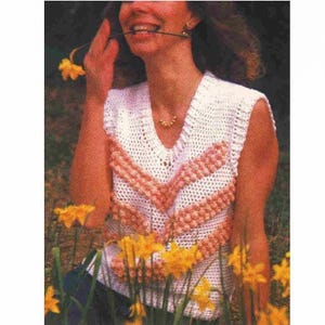 Peut inclure: Un gilet crochet blanc avec un col en V et des accents en chevrons pêche. Le haut sans manches est porté par une personne tenant une fleur jaune. Le fond présente des fleurs jaunes.