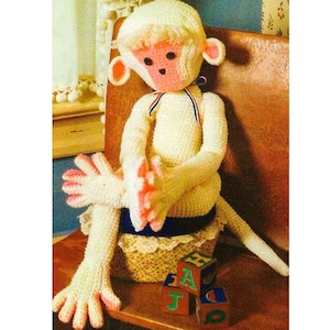 May include: A crocheted monkey doll sitting on a wooden chair with a basket and alphabet blocks. The monkey is wearing a white crocheted outfit with pink accents and a blue and white striped ribbon around its neck.