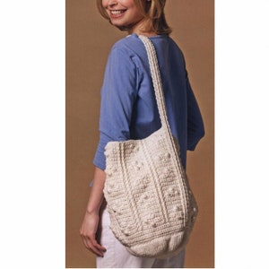 May include: A white crocheted tote bag with a textured design and a long strap. The bag is worn over the shoulder of a person wearing a blue shirt and white pants.