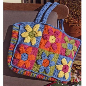 Op de afbeelding: Een kleurrijke, handgehaakte tote bag. De tas heeft een patchwork ontwerp met grote, driedimensionale bloemenapplicaties in geel, oranje, roze en blauw. Het heeft een blauwe rand en bijpassende handvatten.