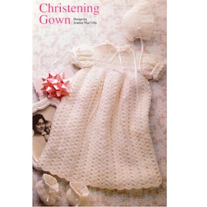 Puede incluir: Un vestido de bautizo de ganchillo color crema con gorro y patucos a juego. El vestido presenta un patrón delicado, mangas cortas y un lazo de cinta. La imagen también incluye una fotografía enmarcada y un lazo rosa.