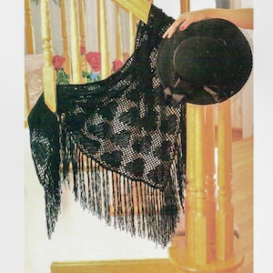 Puede incluir: Un chal negro de ganchillo con un patrón floral y flecos está drapeado sobre una barandilla de madera. Un sombrero negro con una cinta y velo se sostiene cerca. El fondo incluye un vistazo de rosas rojas.