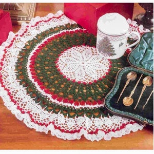 May include: A white crocheted doily with red and green trim. The doily is round and has a scalloped edge. It is placed on a wooden table with a red box and a white teacup with a green tree design.