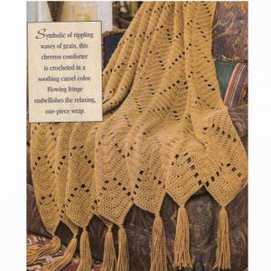 May include: A crocheted chevron throw blanket in a camel color with a fringe border. The blanket is draped over a chair with a patterned fabric.
