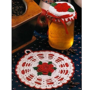 May include: A glass jar filled with a golden liquid, topped with a white crocheted lid adorned with a red rose and green leaves. A matching crocheted doily with a red rose design sits below. Coffee beans and an apple are also visible.