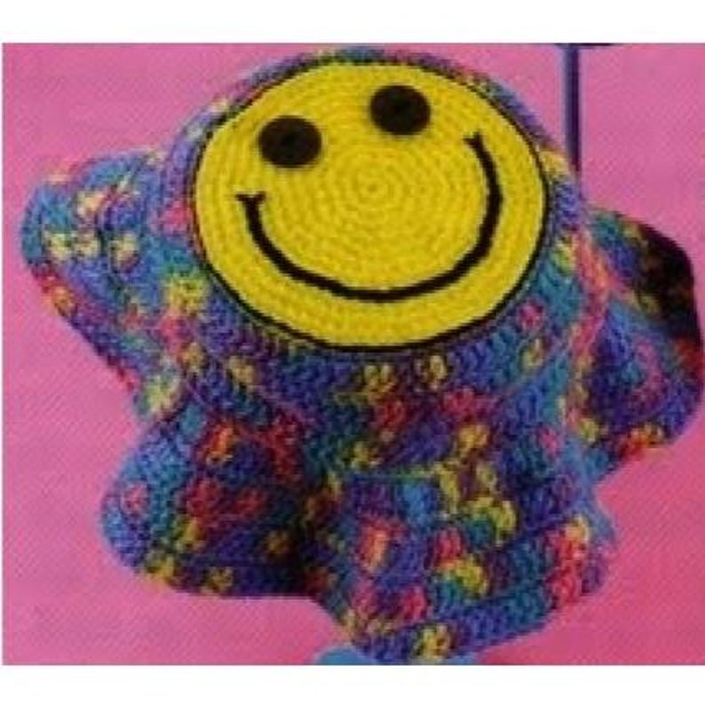 Puede incluir: Un sombrero de ganchillo hecho a mano con una gran carita sonriente amarilla en la parte delantera. El borde y el cuerpo del sombrero presentan un colorido patr&oacute;n moteado de azul, morado, rosa y amarillo. La carita sonriente tiene ojos de bot&oacute;n negros y una boca negra curvada.