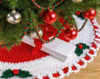 May include: A round, crocheted Christmas tree skirt featuring a vibrant red center, a wide white border, and a scalloped edge decorated with green crocheted holly leaves and small red berries. The skirt is placed beneath a green pine tree.