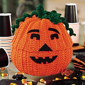 May include: A crocheted orange pumpkin with a green stem and black facial features. The pumpkin has a smiling face with a wide mouth, two eyes, and a triangular nose.