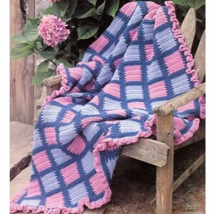 Könnte beinhalten: Eine gehäkelte Decke mit einem karierten Muster in Rosa und Blau. Die Decke hat einen gekräuselten Rand und ist über einen Holzstuhl gelegt.