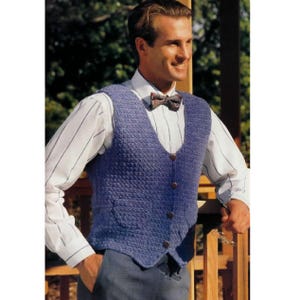 May include: A blue knitted vest with a textured pattern, featuring a V-neck and wooden buttons. The vest is worn over a white dress shirt with thin blue stripes and a patterned bow tie. The person is leaning against a wooden railing.