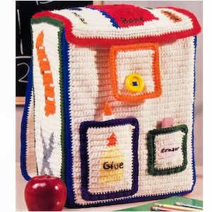 May include: A cream-coloured crochet backpack with blue, red, orange, and green accents. The bag has a flap with the word "Book" and a button closure. It features pockets labelled "Glue" and "Eraser", and a side with an orange pencil design.
