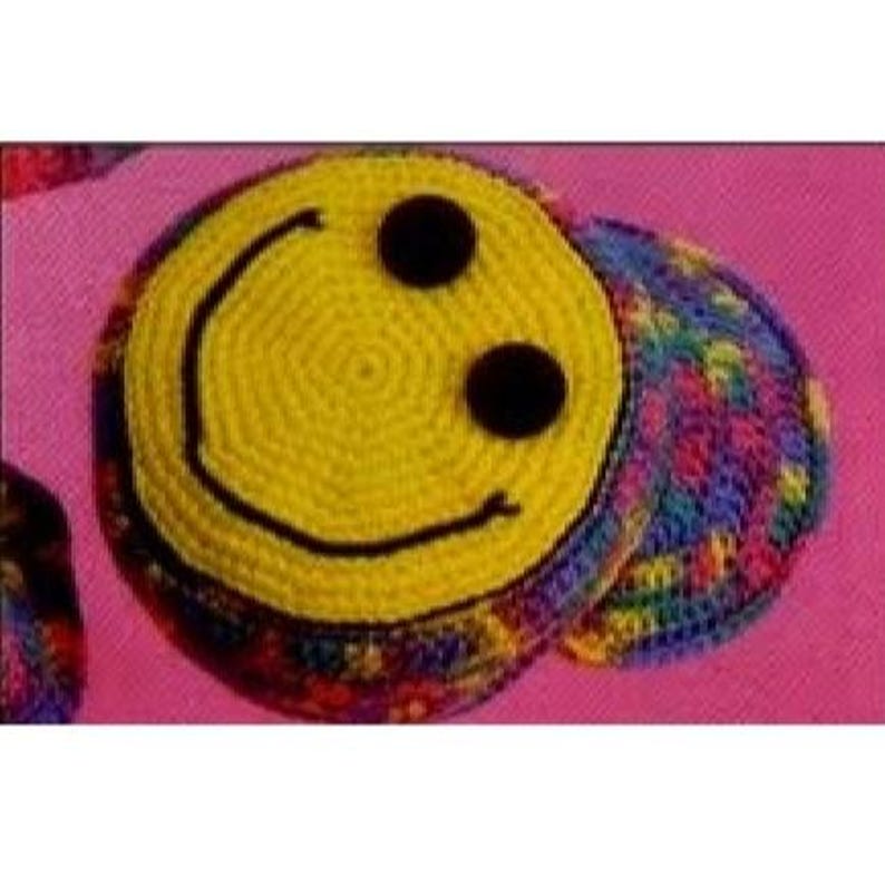 Puede incluir: Posavasos amarillo hecho a mano con una cara sonriente negra. El posavasos tiene dos ojos negros de bot&oacute;n y una boca negra curvada. El borde es una colorida mezcla de hilo arco&iacute;ris. El posavasos est&aacute; sobre una superficie rosa.