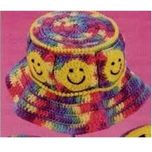 Puede incluir: Un sombrero de pescador de ganchillo colorido con un patr&oacute;n de remolino de arco&iacute;ris en la parte superior y un ala ancha. El sombrero presenta tres caras sonrientes amarillas alrededor de la banda. El sombrero est&aacute; hecho de hilo en tonos de rojo, amarillo, azul y morado.